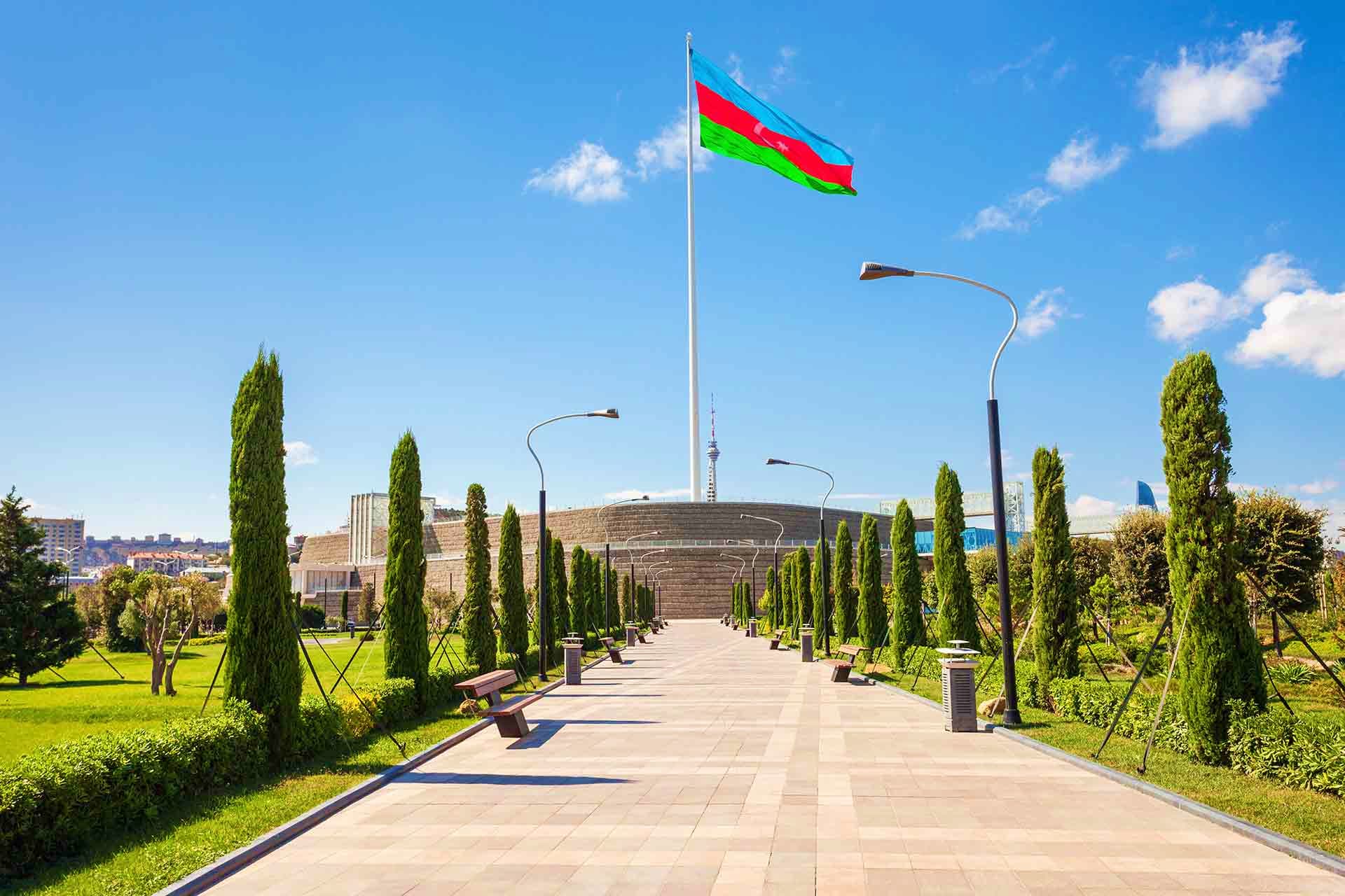 Azerbaijan Visa Azerbaijan flag flying above a modern pavilion building in Baku with cypress trees lining a paved pathway and city skyline in